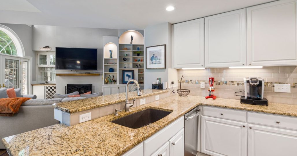 A modern kitchen with granite countertops opens into a stylish living room, featuring elegant furniture and built-in shelves.